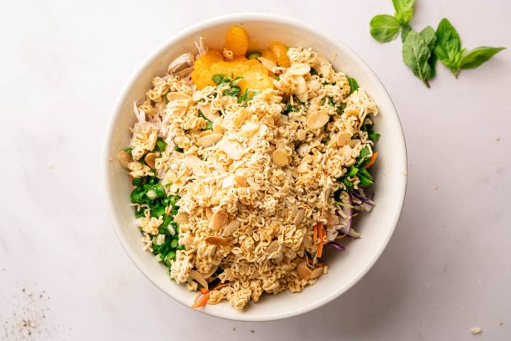 A bowl of Asian-inspired salad with crunchy ramen noodles, shredded vegetables, mandarin oranges, and sliced almonds on a light surface with basil leaves.